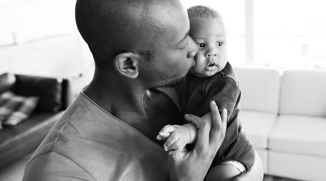 Young afro-american father at home holding his cute baby son in his arms, kissing him on cheek.