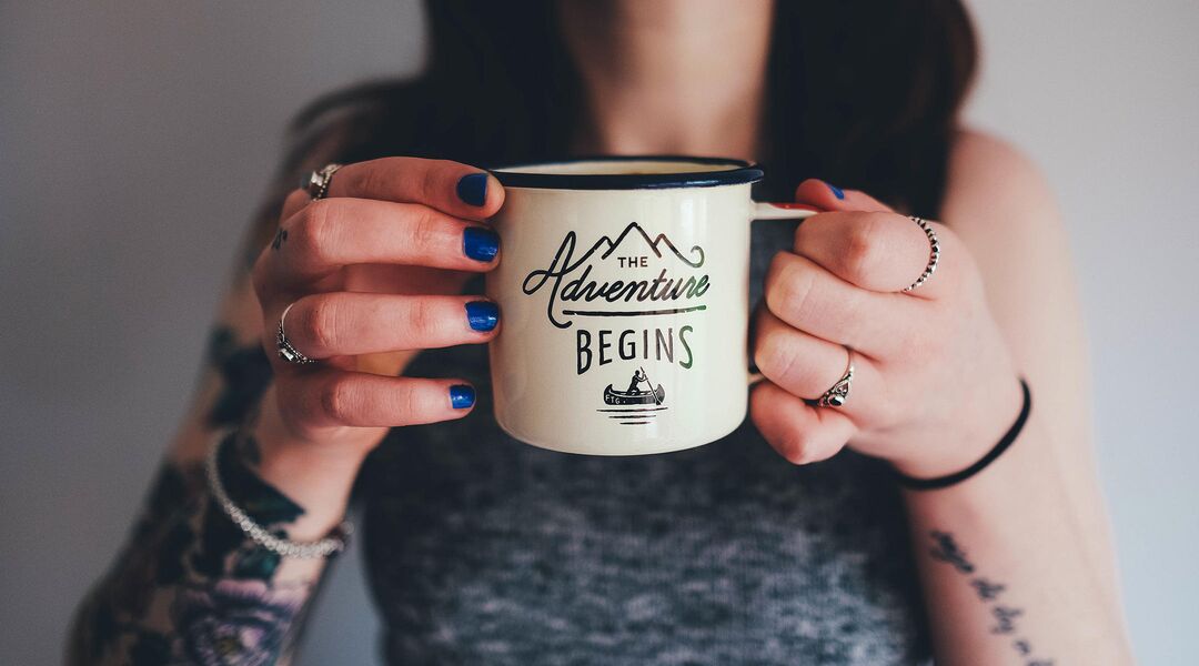 Close up of alternative woman with blue nail polish holding mug that says: the adventures begin. 