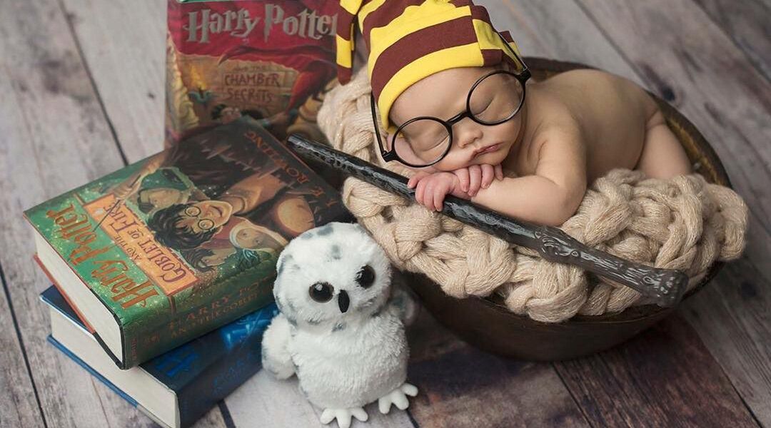 A baby sleeps with a Gryffindor sleeping cap and wand in hand, next to three Harry Potter books and a stuffed Hedwig owl.
