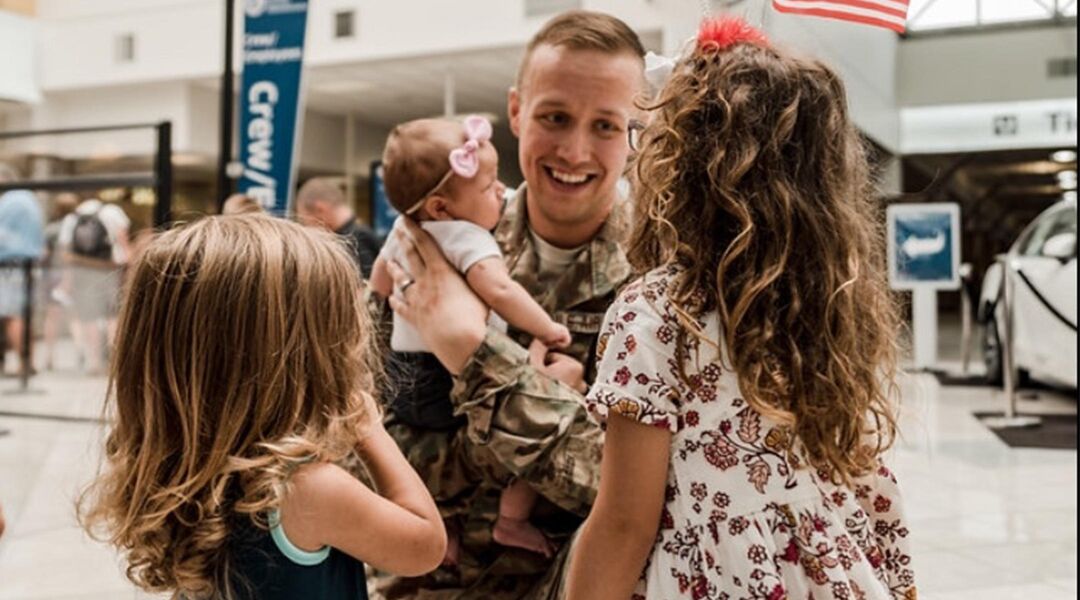Military dad holding new baby while daughters stand in front