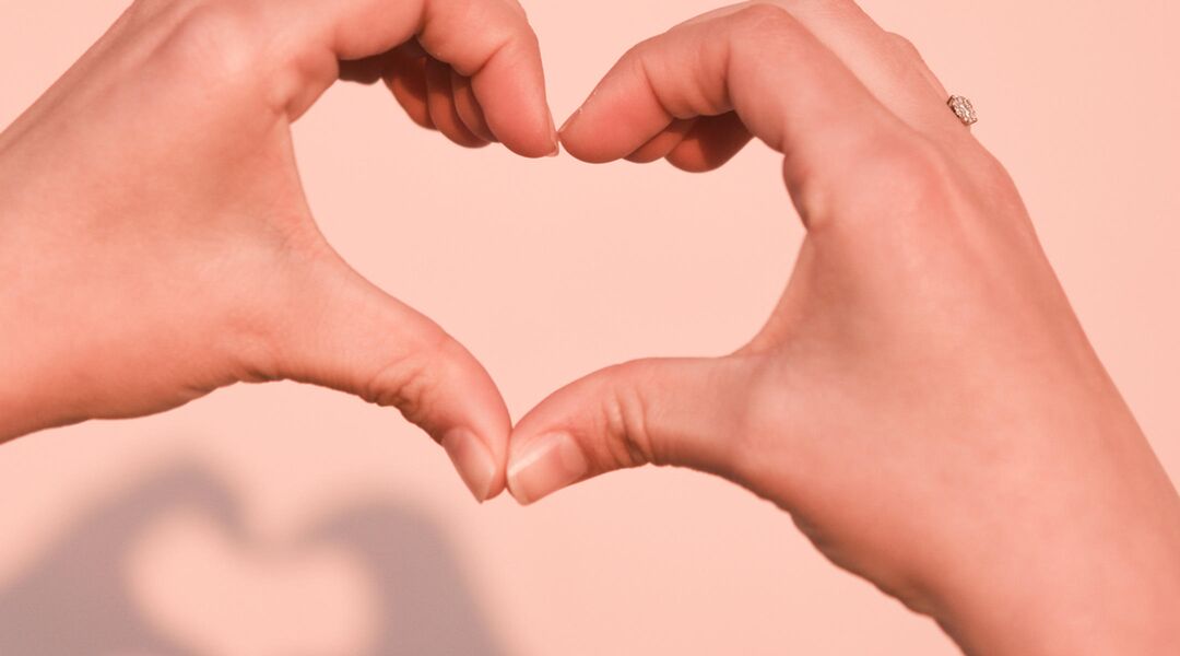 Two hands forming heart shape against pink background.