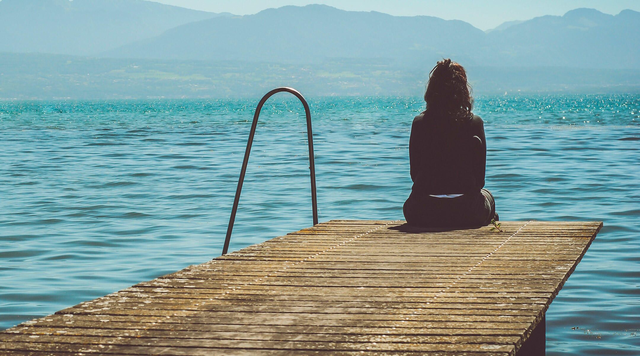 sad woman sitting on dock surrounded by beautiful lake and mountain landscape