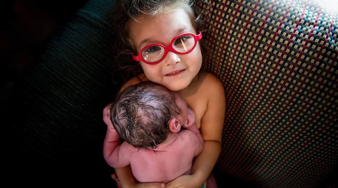 Little girl with glasses holding newborn sibling skin-to-skin