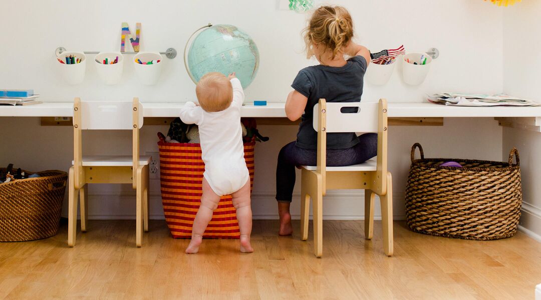 toddler standing next to young child seated at desk