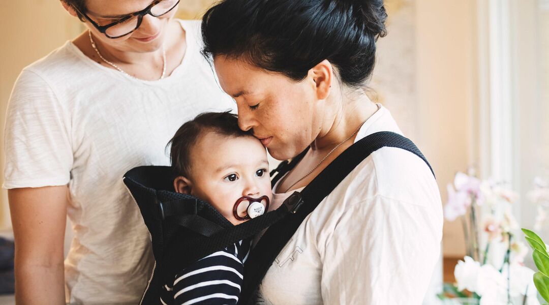 Same sex couple mom kissing baby in baby carrier. 