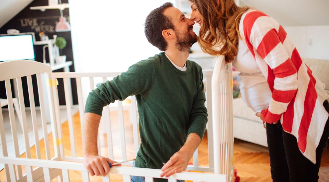 Pregnant woman affectionately rubbing noses with her partner who is building a baby crib.
